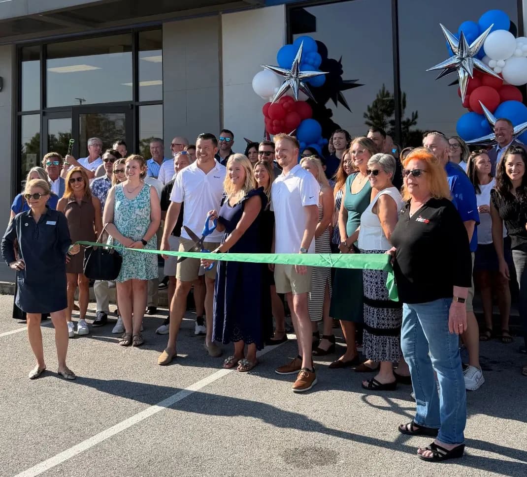 Crowd gathered outside the Team 1 building for a chamber ribbon cutting, with a green ribbon stretched across the front row