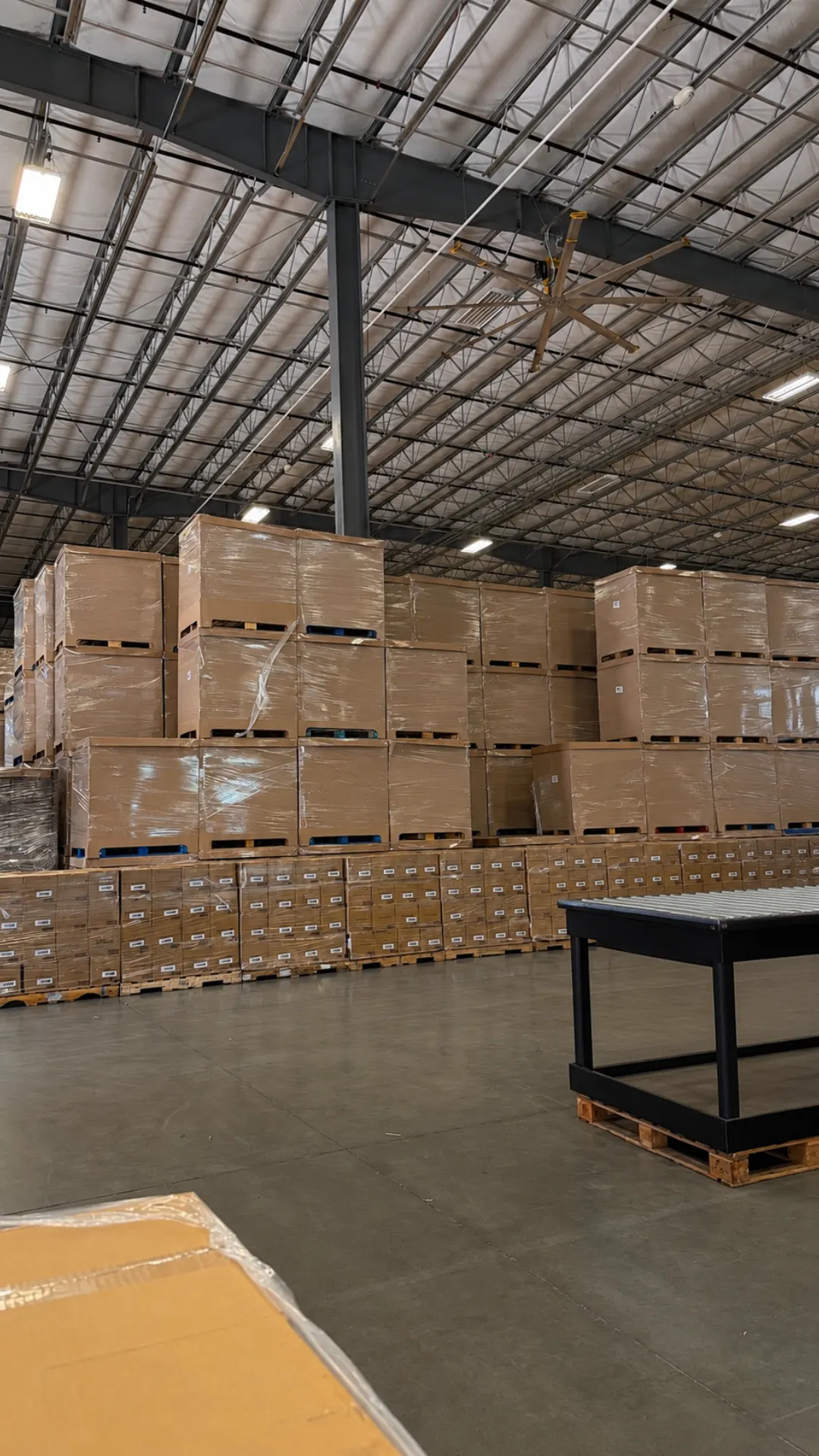 Shrink-wrapped pallets floor-stacked three high under a warehouse ceiling fan